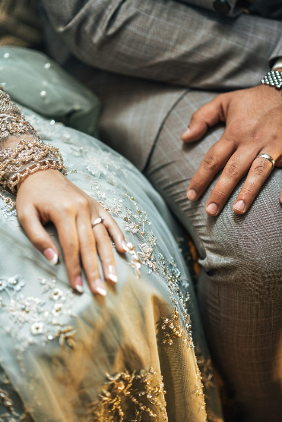 A bride and groom seated side by side, her hand resting on her embroidered teal lehenga, his hand resting on his patterned grey trousers; both wearing wedding rings, hands close but not touching.