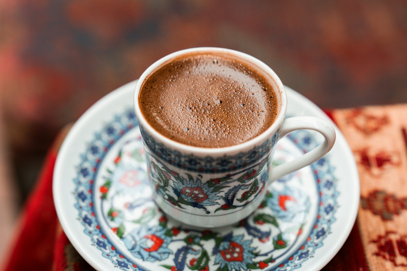 A small porcelain cup of Turkish coffee on a saucer painted with Iznik-style florals, set on a warm woven rug.