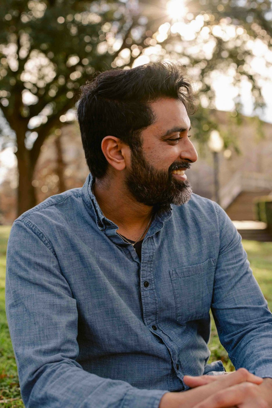 A man in his thirties seated outdoors in soft natural sunlight, looking off to the side with a quiet, easy smile.