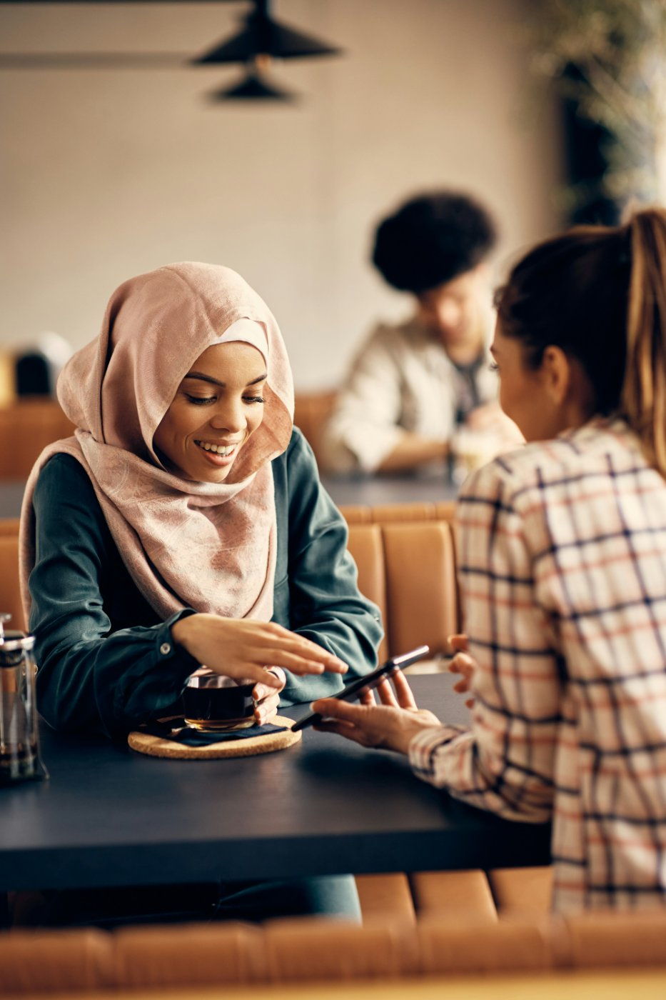Two friends in conversation over tea, one in a soft pink hijab, sharing a quiet exchange.